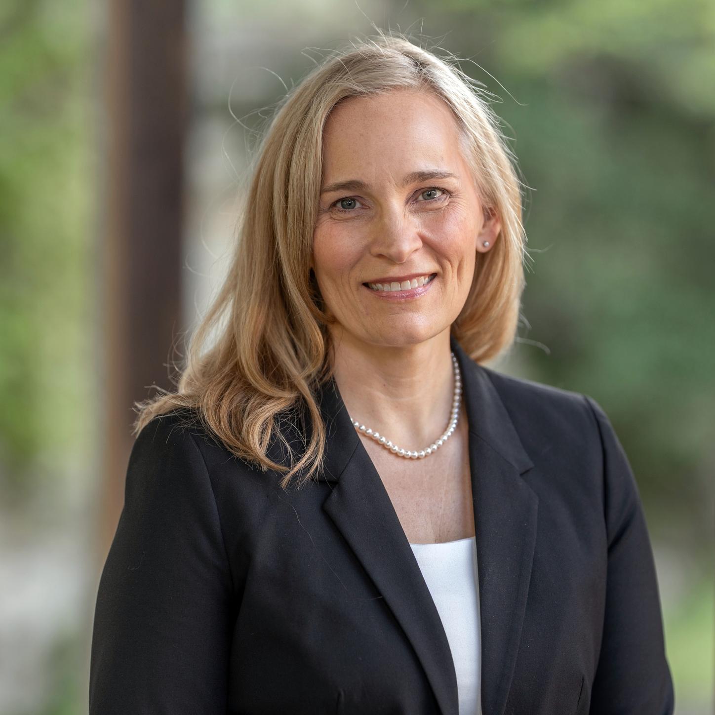 Professional portrait of Elaine Becraft, Chief Human Resources Officer (CHRO), with shoulder-length hair, wearing a dark blazer, white top, and pearl necklace, against a softly blurred green background.