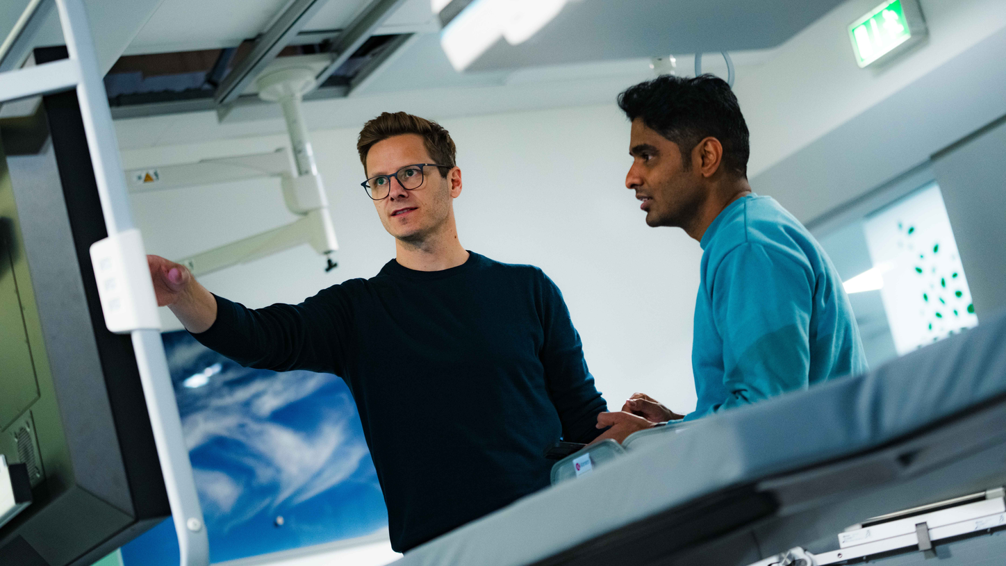 Zwei Personen stehen neben medizinischer Bildgebungstechnik in einem Testlabor. Eine Person zeigt auf einen großen Monitor und erklärt etwas.