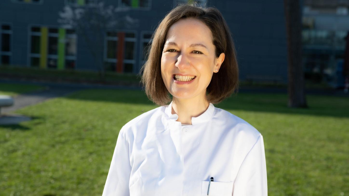 Medium close-up portrait photo of Ulrike Attenberger wearing half-long, dark brown hair, a doctor's white lab coat and smiling into the camera. A blurred image of the Bonn University Hospital building is visible in the background.