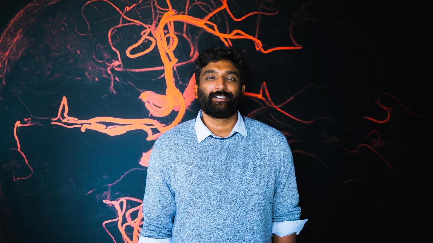 Portrait of Ullaskrishnan Poikavila against a dark wall with orange graphic lines that look like brain cells.