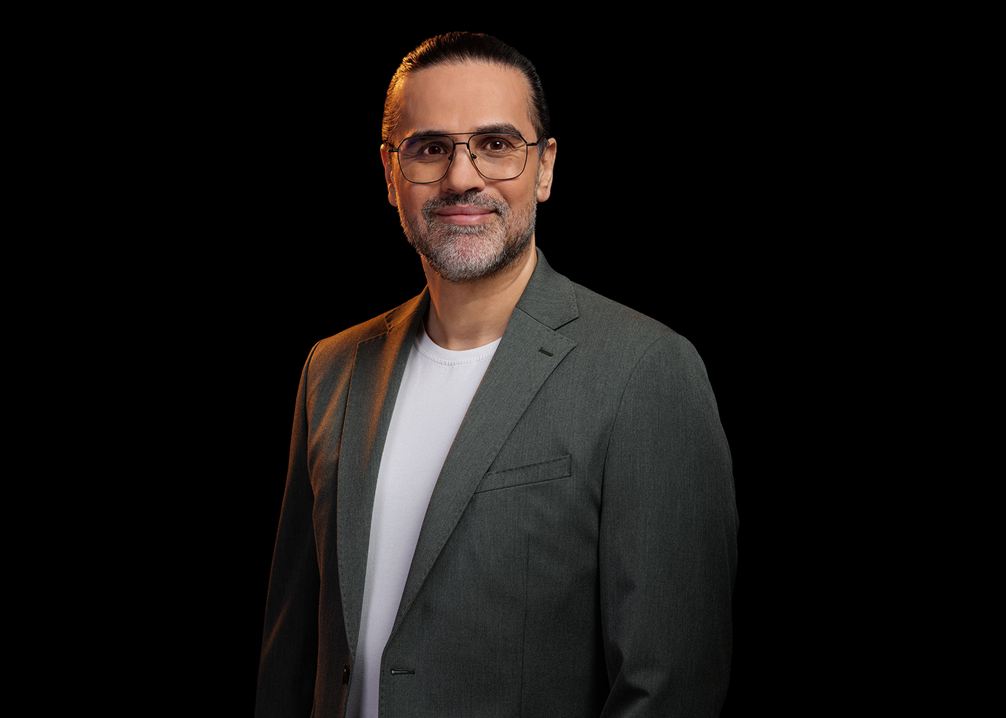 Studio portrait of a person wearing glasses, a gray blazer, and a white shirt, standing against a black background.