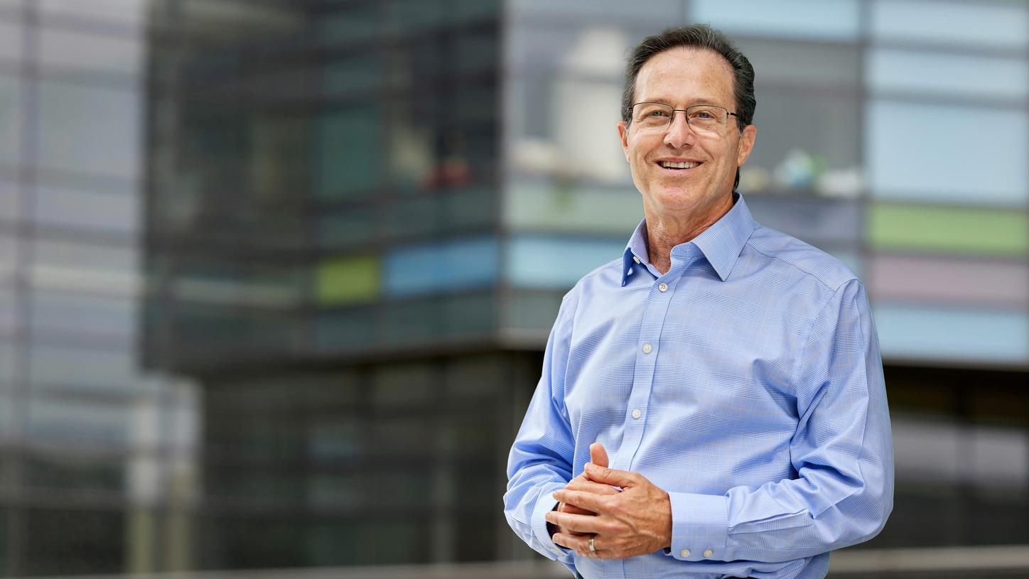 Person wearing a light blue button-down shirt stands with hands loosely clasped in front, positioned outdoors against a modern glass building with horizontal architectural lines.