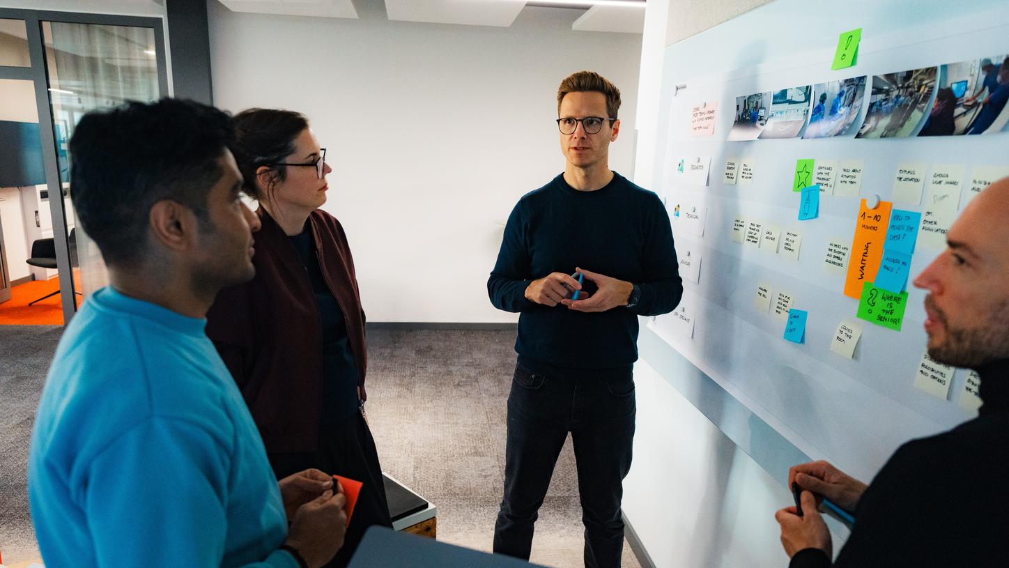Eine kleine Gruppe von Kolleg:innen steht in einem modernen Büro um ein Whiteboard. Eine Person, Timo Röhrig, hält einen Marker und gestikuliert, während er die Diskussion leitet. Auf dem Whiteboard sind Notizen, Skizzen und Haftnotizen zu sehen; die anderen hören zu und halten Stifte oder Notizblöcke.