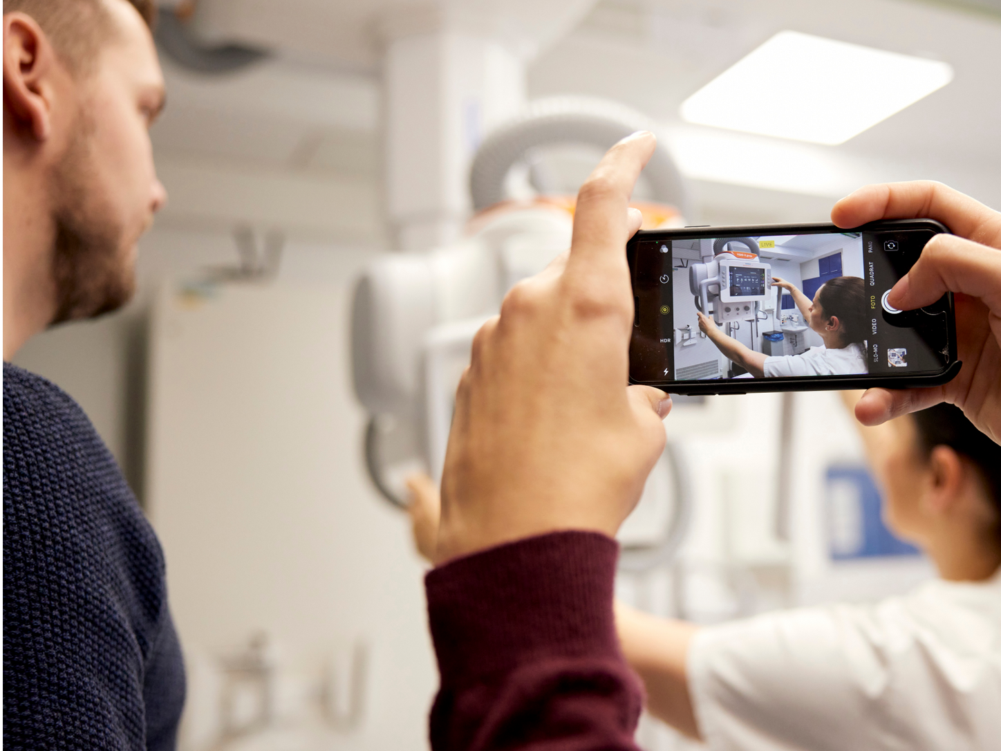 Ein Mann hält ein Handy im Vordergrund und macht ein Foto von einem Gesundheitsarbeiter, der eine Maschine benutzt.
