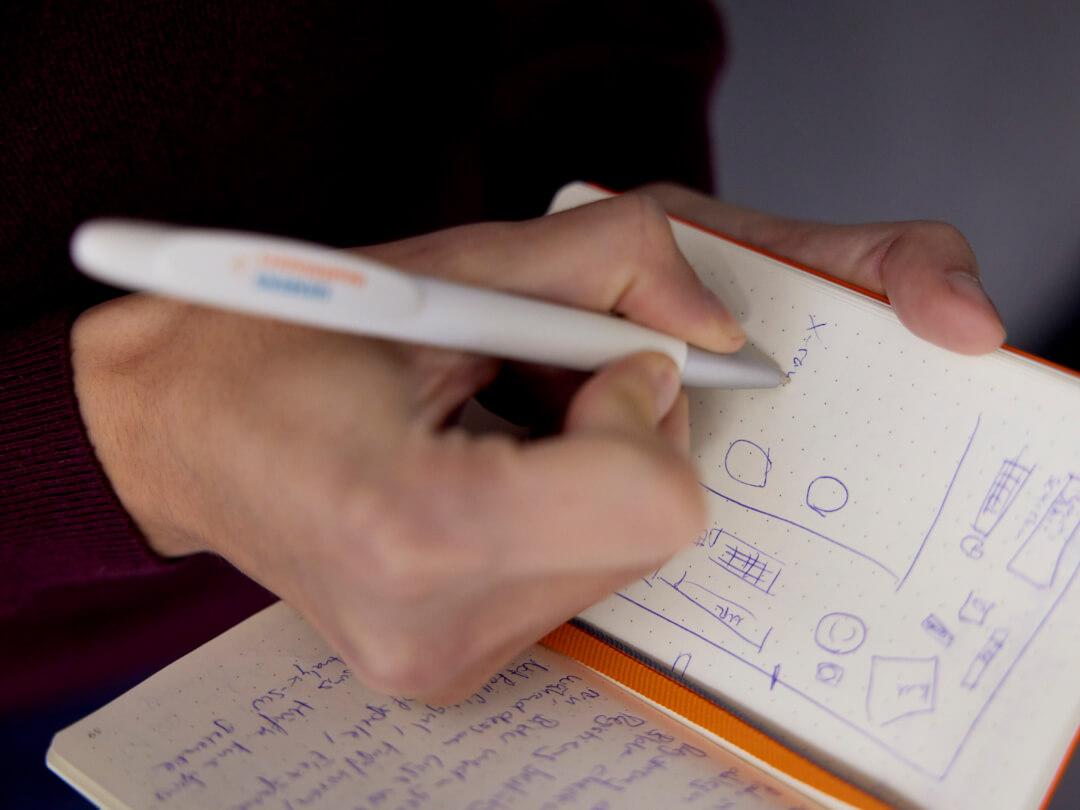 Eine Person, die Notizen in einem orangefarbenen Notizbuch mit weißen Seiten macht, mit einer Siemens Healthineers-Seite.