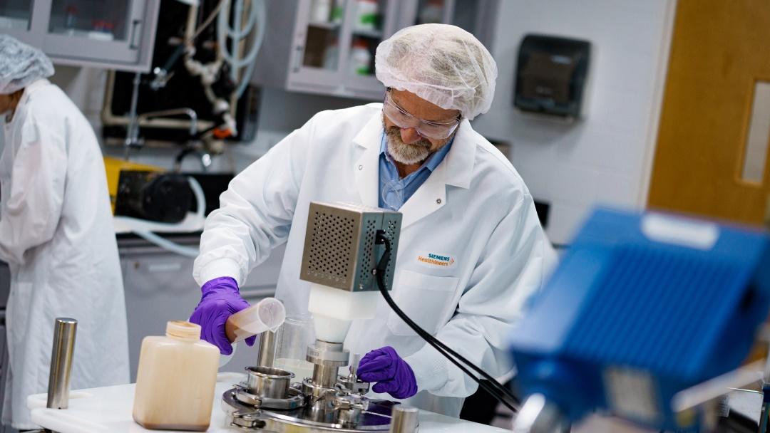 Jim Freeman im Labor mit Laborkittel, Schutzbrille und Haarnetz. Er schütet eine braune Flüssigkeit in einen großen Behälter.