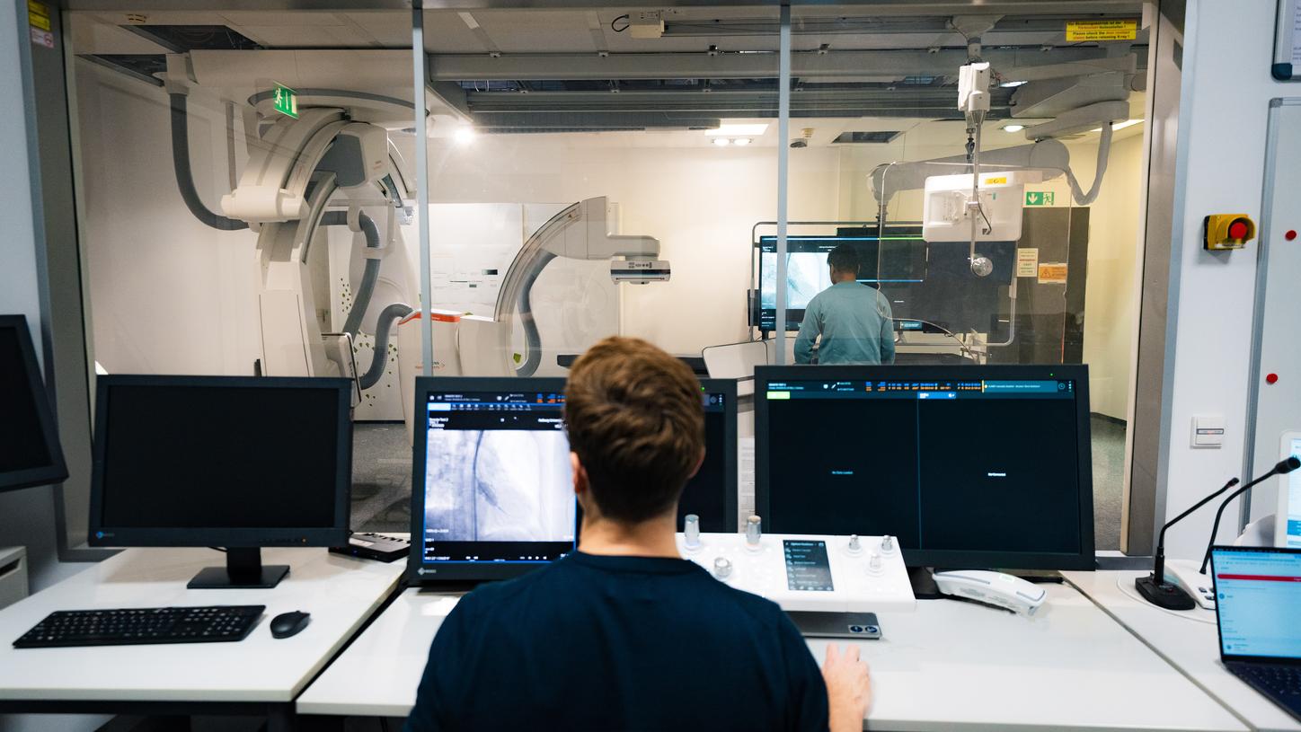 Eine Person sitzt an einem Leitstand mit mehreren Monitoren, auf denen medizinische Bilddaten angezeigt werden. Durch ein großes Fenster blickt sie in einen angrenzenden Raum, in dem eine weitere Person in einer klinischen Umgebung einen Prototyp testet.