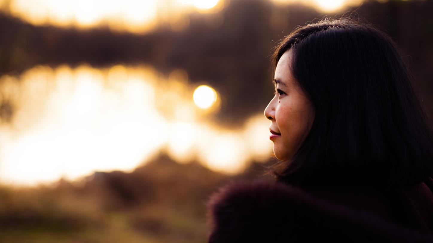 Photograph portrait of from Ren-Yi Lo taken from behind her. She stands on the shore of a lake, looking to her left, smiling. 