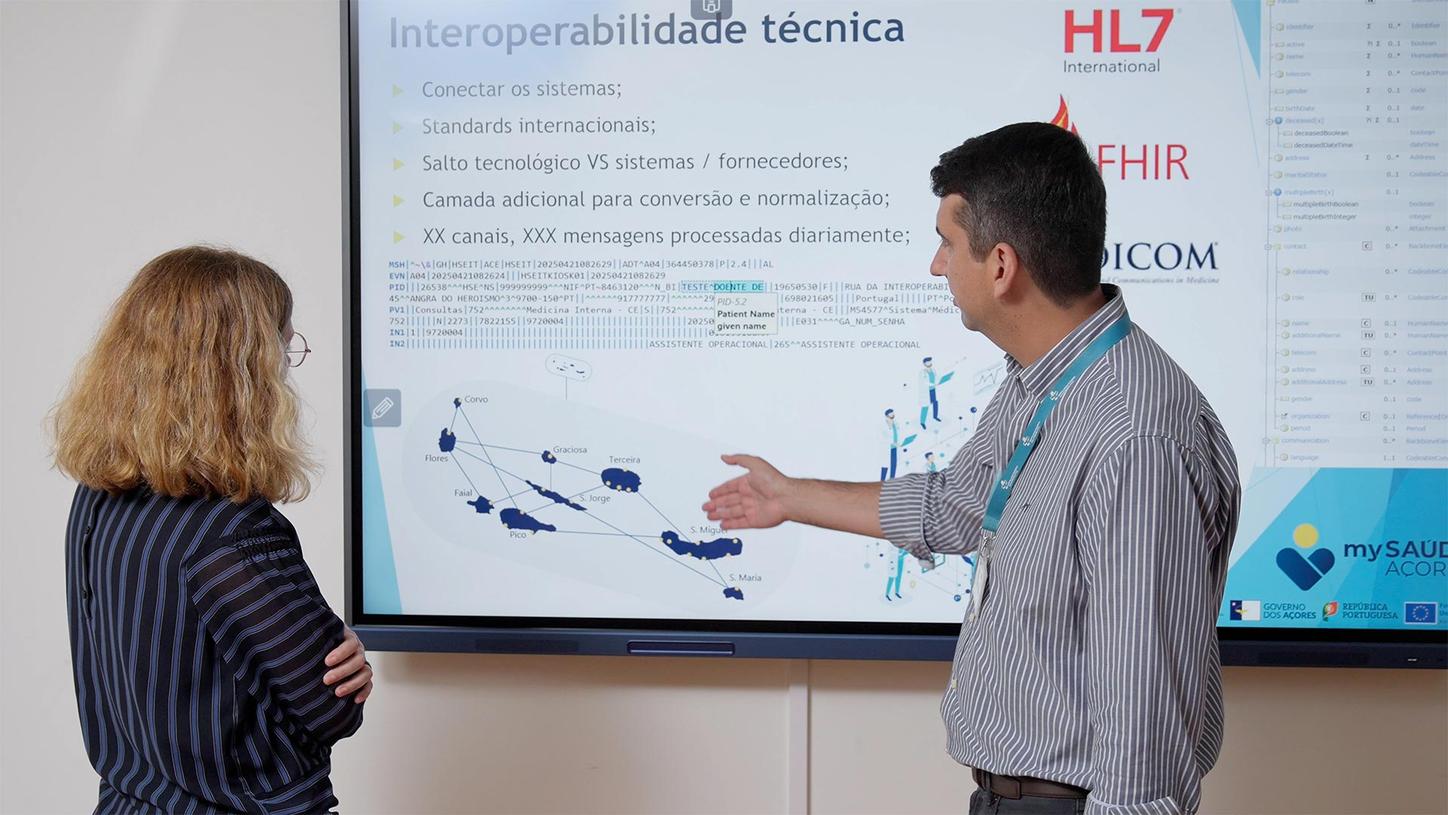 Two people in front of a large screen, one pointing at a presentation slide with technical text and a diagram.