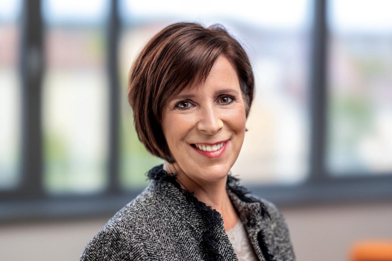 Darleen Caron mit kurzem Haar und strukturierter Jacke vor einem weich verschwommenen Bürohintergrund.