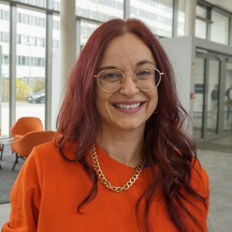 Portrait of Corporate Journalist Katja Gäbelein in the reception area of a Siemens Healthineers building. She is wearing an orange top and looking at the camera.