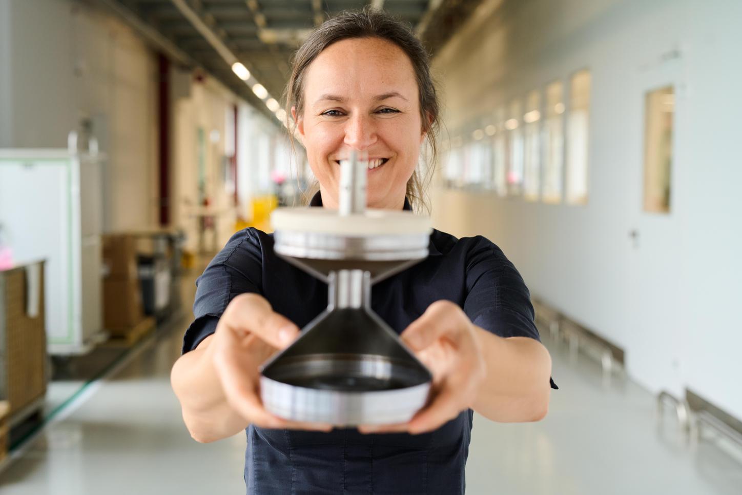 The photo shows Anja Fritzler in a dark blouse. She holds an X-ray tube in her hand as if it were a dumbbell and smiles at the camera. In the background, an office environment is blurred.  