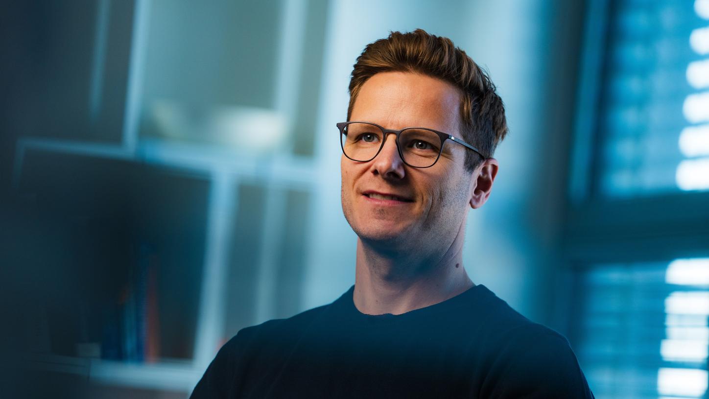 Timo seated indoors, wearing a dark shirt with soft blue lighting and a blurred office background.