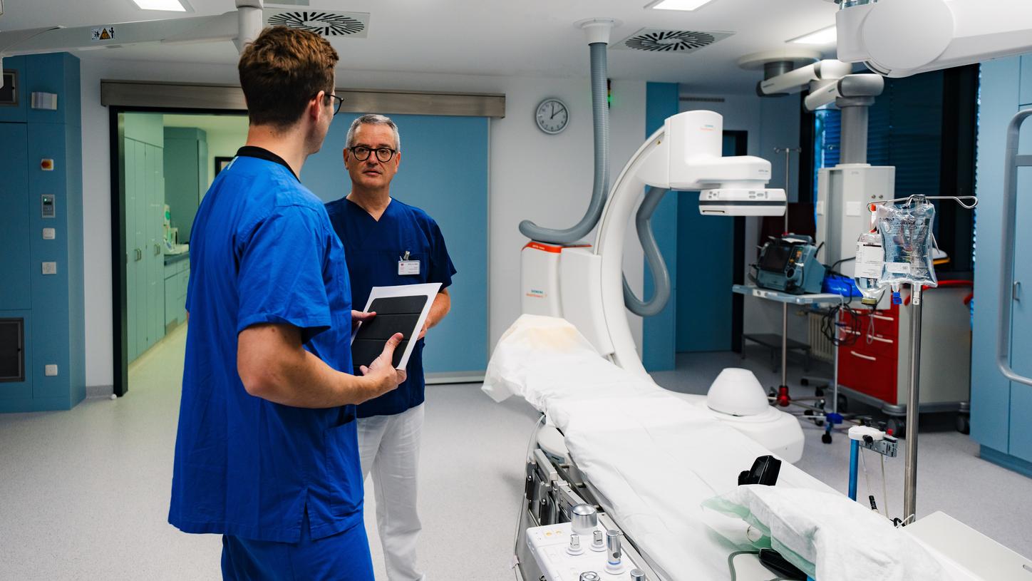 Zwei Personen besprechen Arbeitsabläufe anhand eines Tablets in einem Herzkatheterlabor mit moderner Bildgebungstechnologie.