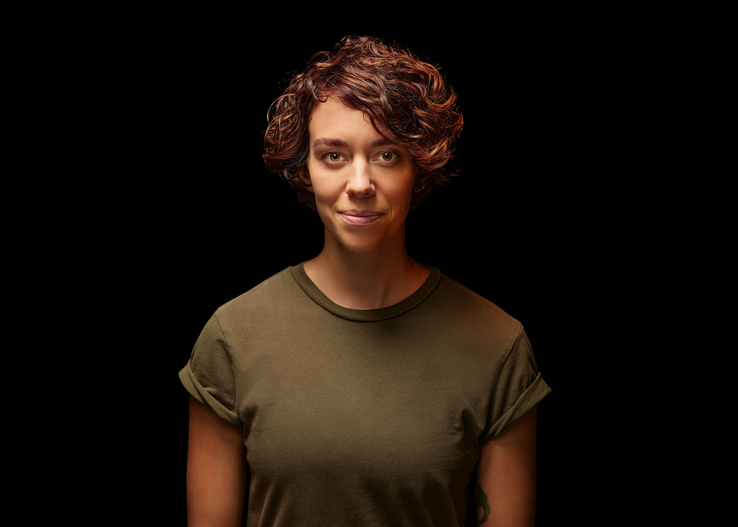 Studio portrait of a person with short, curly hair, wearing a plain short‑sleeve shirt, facing the camera against a black background.