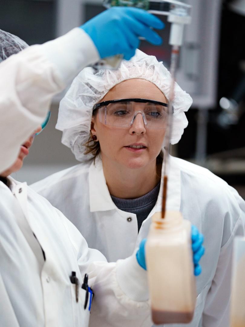 Zwei Kolleginnen im Labor mit Laborkittel, Schutzbrille und Haarnetz. Sie entnehmen eine braune Flüssigkeit mit einem Messbehälter.