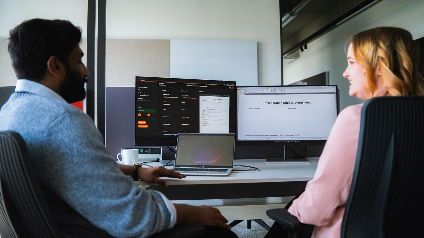 Ullaskrishnan Poikavila and a colleague work together at a computer workstation, reviewing information displayed on multiple screens.