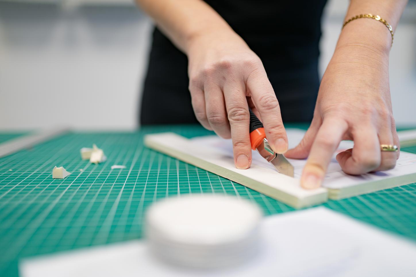 Close-up of hands cutting a thin sheet of material with a craft knife on a cutting mat.