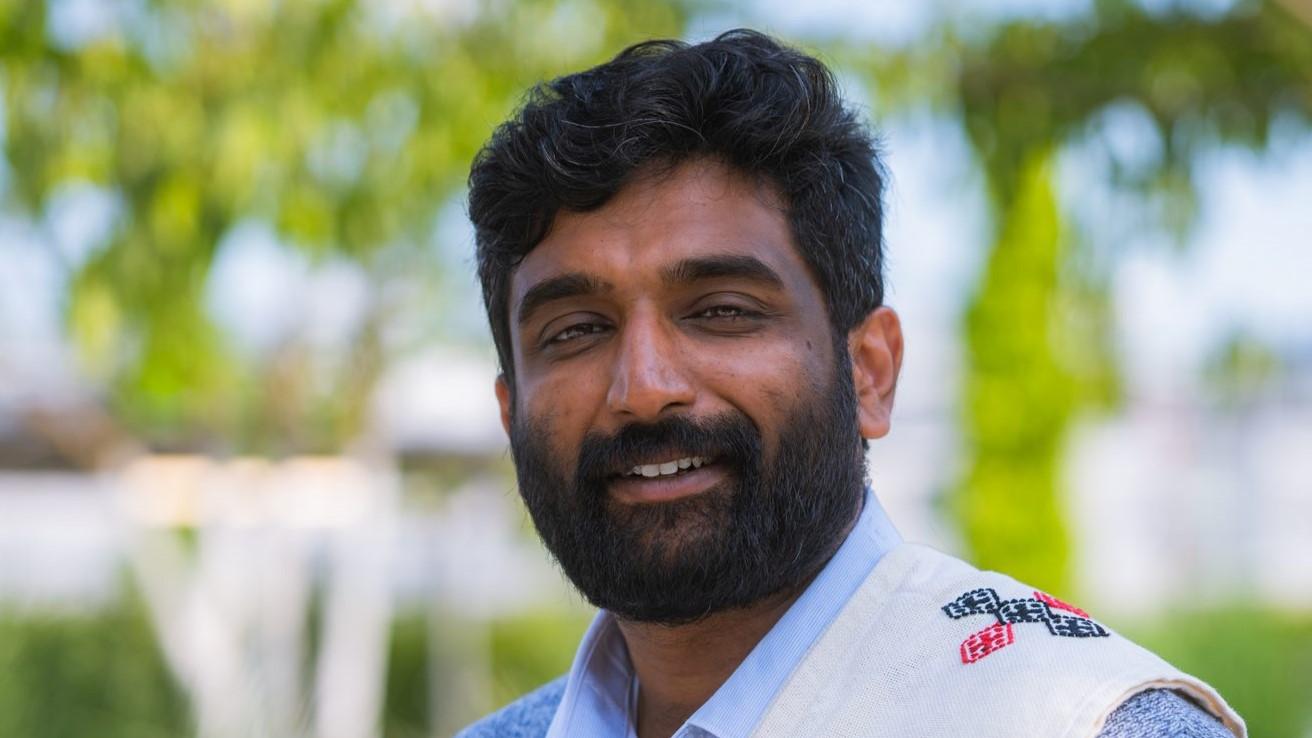 Portrait of Ullas Poikavila. He is wearing a traditional Poothkuli shawl from the Toda community draped over his shoulder and is smiling at the camera.