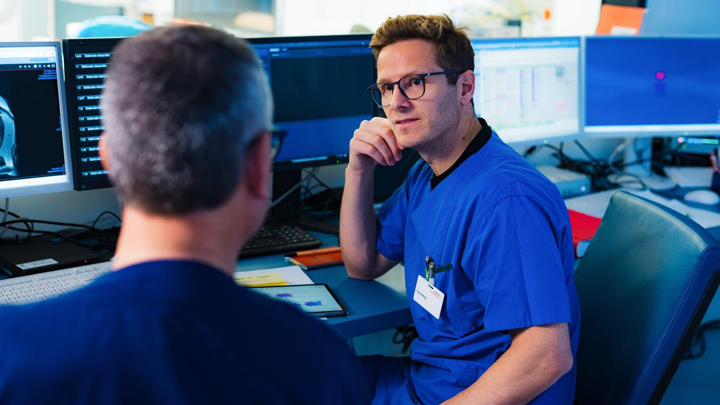 Eine Person in blauer OP‑Kleidung sitzt an einem Arbeitsplatz und bespricht etwas mit einer klinischen Fachkraft. Mehrere Bildschirme zeigen klinische Informationen in einer Leitstellen‑ähnlichen Umgebung.