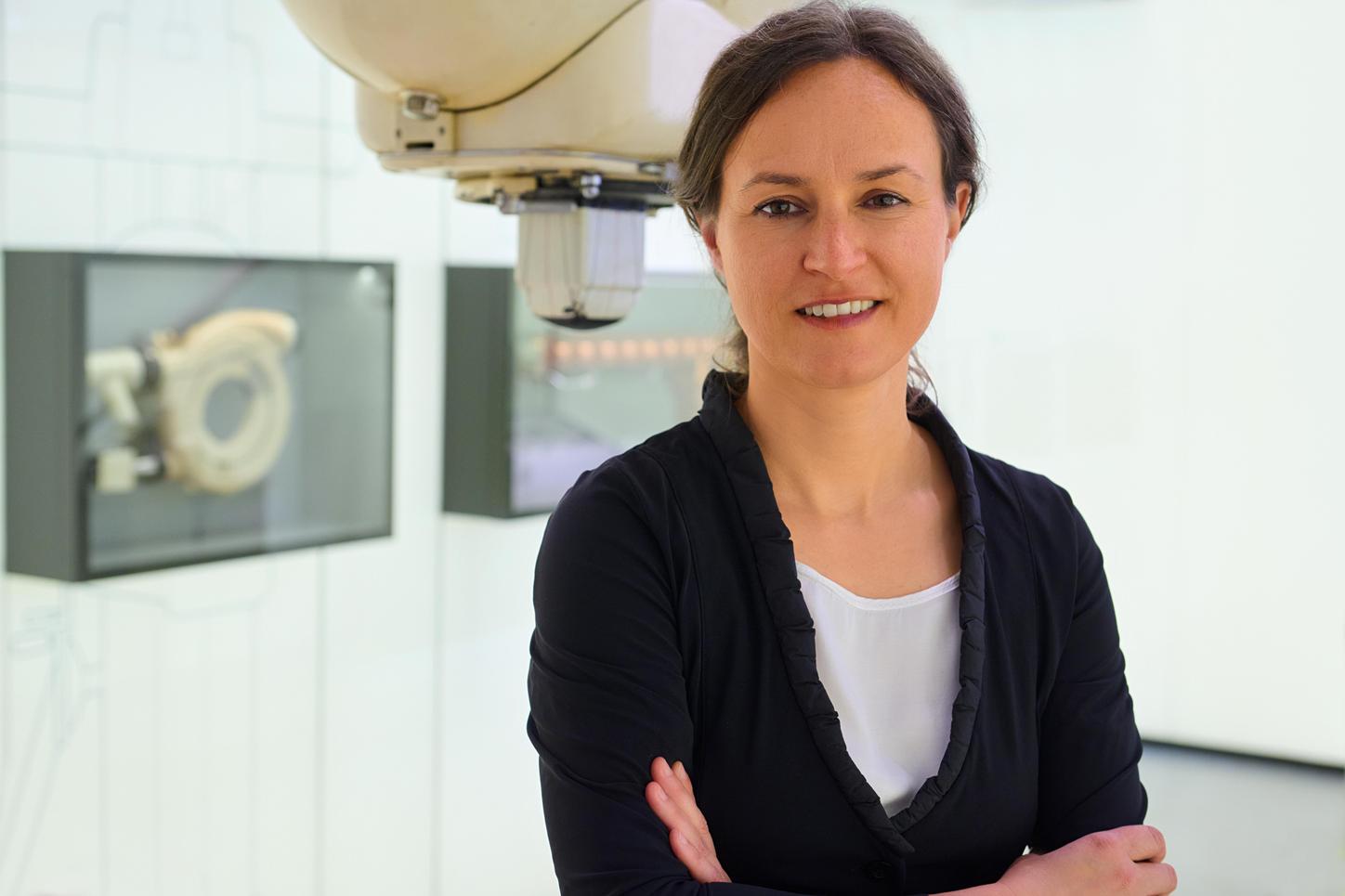 Portrait photo of Anja Fritzler. She wears a black blouse, her hair is tied in a ponytail. She is standing in the MedMuseum of Siemens Healthineers, with her hands clasped in front of her chest and smiling towards the camera. In the background you can see historical medical devices.  