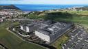 Aerial view of a large modern building complex with parking lots and surrounding fields near a coastal town