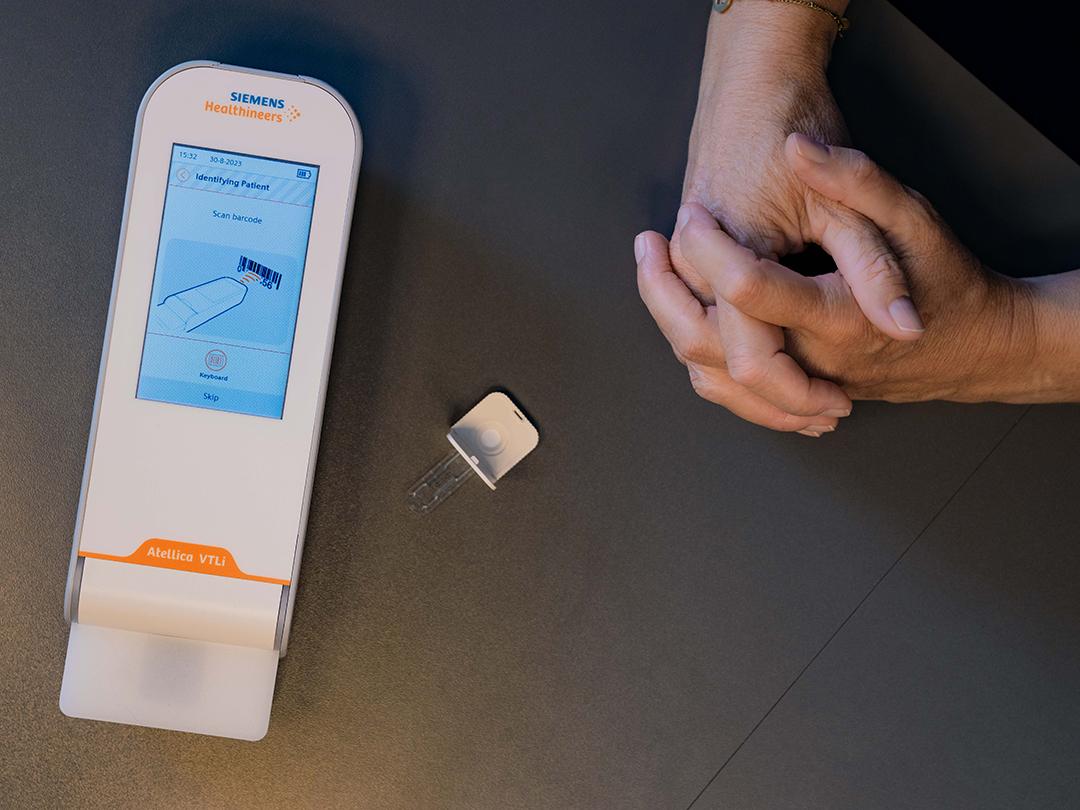 Top shot showing the handheld device, Atellica VTLi, and an open cartridge, on a desk. Femke's hands are shown as well.