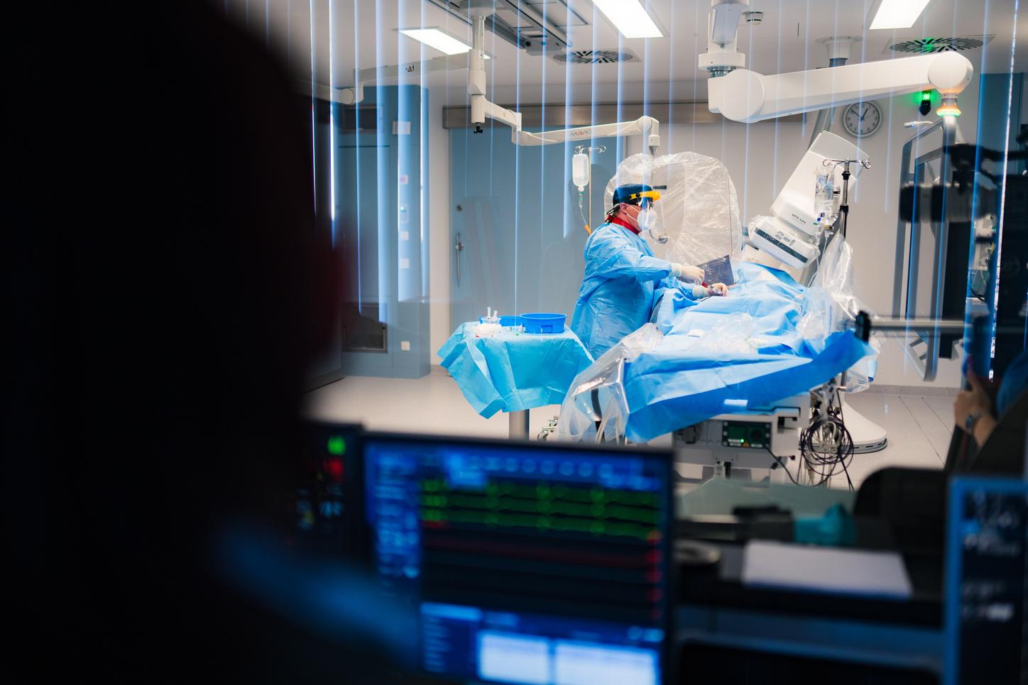 Eine medizinische Fachkraft in Schutzkleidung führt in einem Katheterlabor einen interventionellen Eingriff durch. Der Eingriff ist durch das Fenster des Kontrollraums zu sehen, im Vordergrund sind Überwachungsmonitore sichtbar.